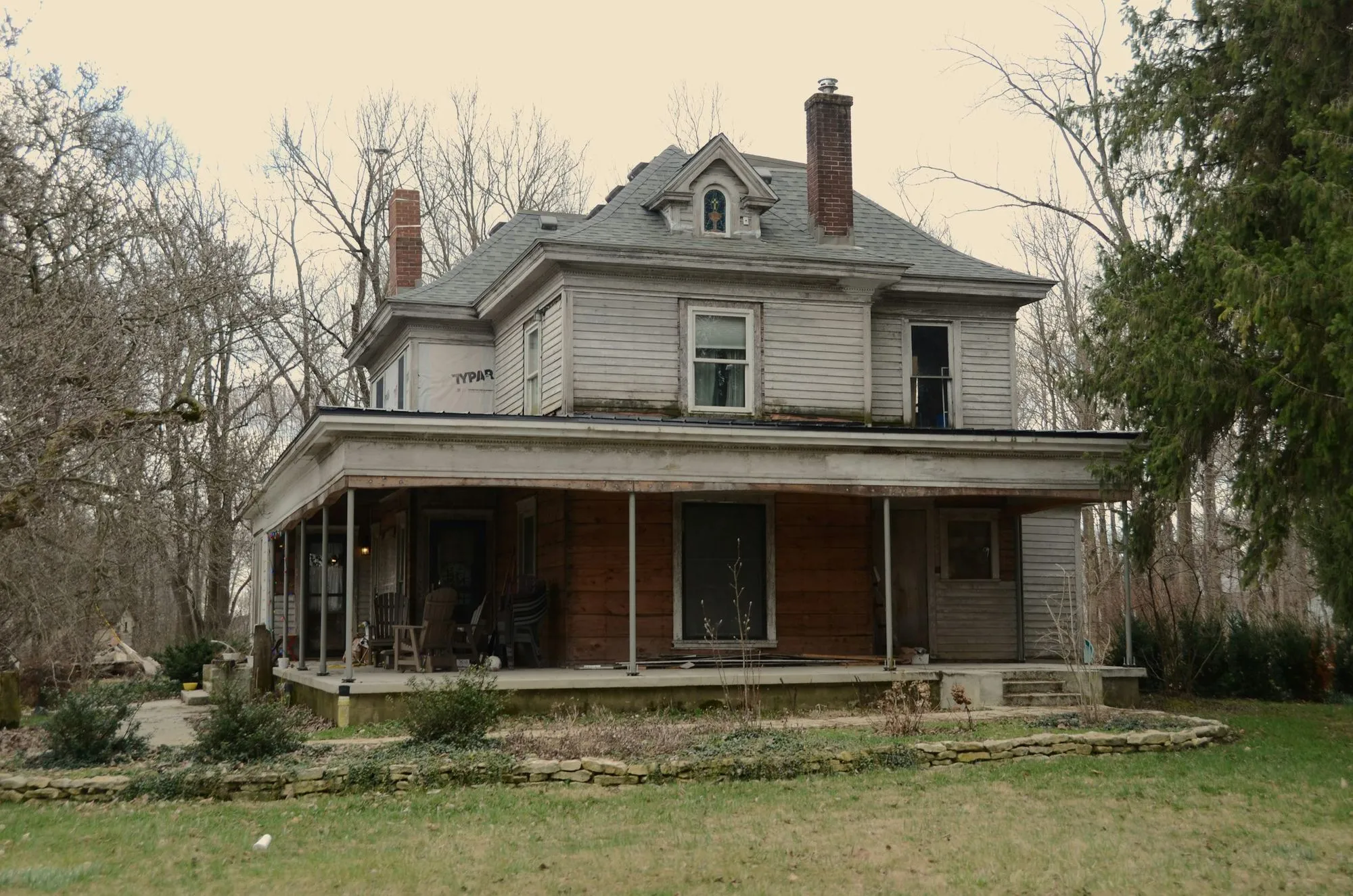 A weathered two storey house with a wraparound porch and a chimney, bare trees behind